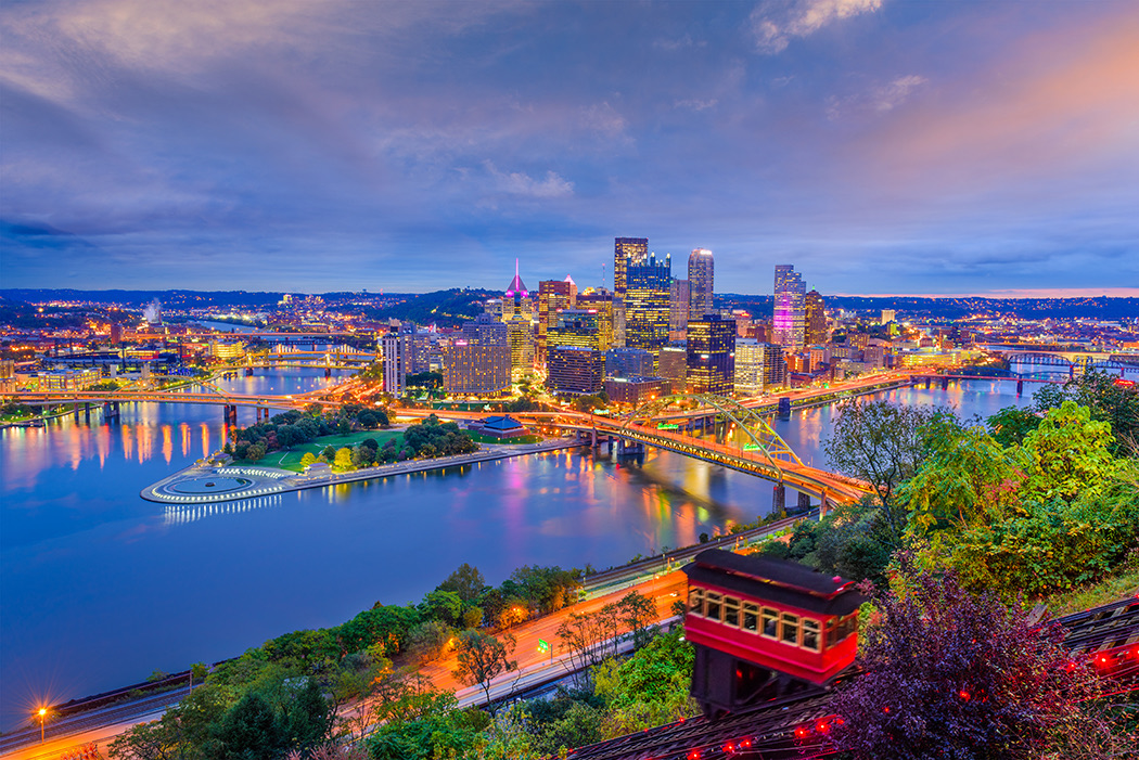Downtown Pittsburgh at dusk from Mount Washington — the skyline and three rivers lit up, with an incline car descending in the foreground.