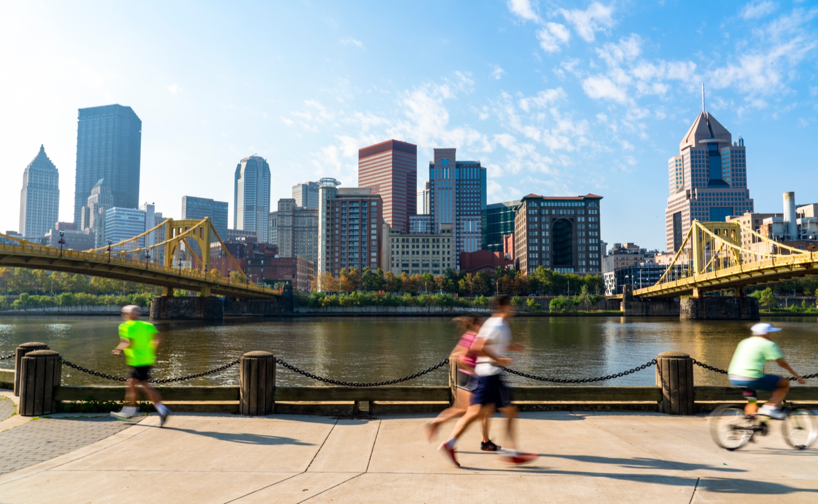 A sunny Saturday on Pittsburgh's North Shore riverfront — joggers and a cyclist along the Allegheny, the two yellow bridges, and the downtown skyline across the water.
